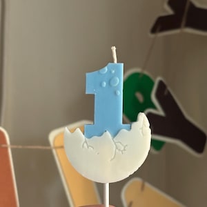 K&ouml;nnte beinhalten: Eine hellblaue Kerze in Form der Zahl Eins, die aus einer wei&szlig;en, rissigen Eierschalenbasis hervorgeht. Die Kerze hat einen kleinen wei&szlig;en Docht an der Spitze und ist an einem d&uuml;nnen wei&szlig;en Stab befestigt, der als dekorativer Aufsatz dient.