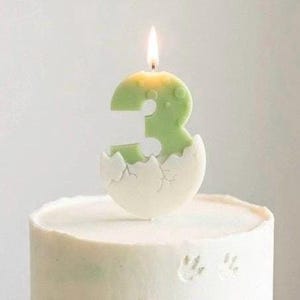 K&ouml;nnte beinhalten: Eine hellgr&uuml;ne Geburtstagskerze in Form der Zahl Drei, die einen aus einer wei&szlig;en, rissigen Eierschale schl&uuml;pfenden Dinosaurier darstellt. Die Kerze brennt mit einer kleinen Flamme und steht auf einem wei&szlig; glasierten Kuchen mit Textur.