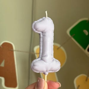 May include: A light purple candle shaped like the number one, featuring a textured, inflated balloon-like appearance with a visible white wick at the top and a small wooden stick extending from the bottom base for support.