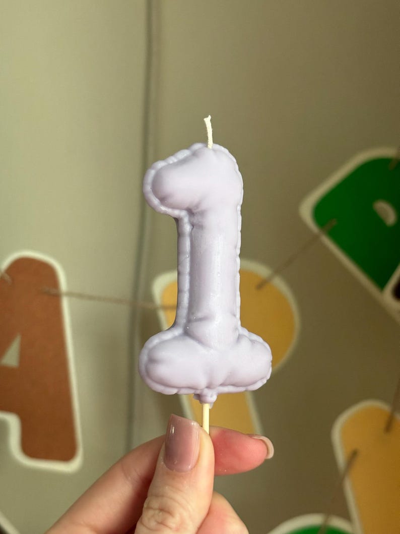 May include: A light purple candle shaped like the number one, featuring a textured, inflated balloon-like appearance with a visible white wick at the top and a small wooden stick extending from the bottom base for support.