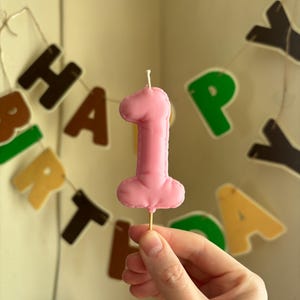 May include: A pink number one shaped candle with a smooth wax finish and a single white wick at the top, mounted on a thin wooden pick. The candle features a rounded, puffy design and is presented against a blurred background of birthday banner letters.