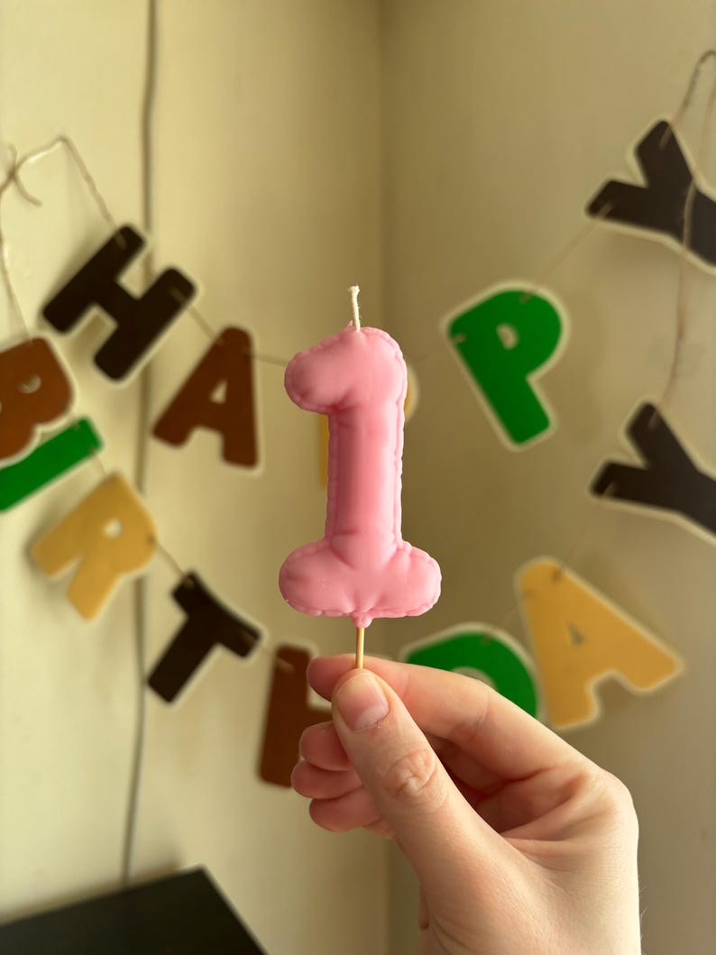 May include: A pink number one shaped candle with a smooth wax finish and a single white wick at the top, mounted on a thin wooden pick. The candle features a rounded, puffy design and is presented against a blurred background of birthday banner letters.