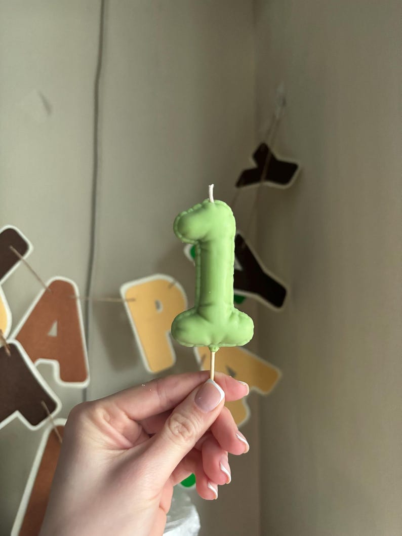 May include: A light green number one shaped candle with a smooth, rounded texture and a visible wick at the top. The candle is attached to a thin, light-colored wooden pick at the base, designed for insertion into a cake or other food item.