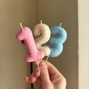 May include: Three number-shaped candles in pink, cream, and light blue. Each candle features a stitched edge detail and a small white wick at the top. The candles are mounted on thin wooden sticks, forming the sequence one, two, and three.