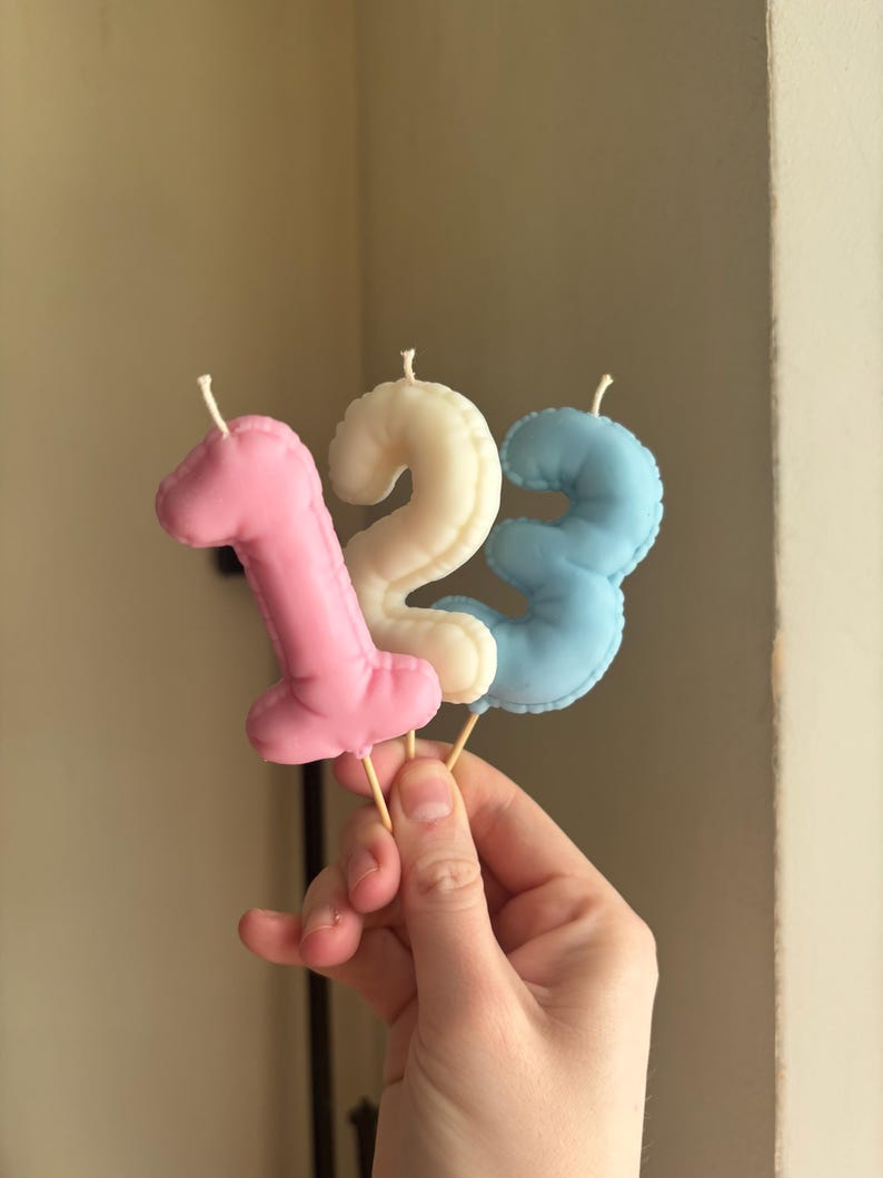 May include: Three number-shaped candles in pink, cream, and light blue. Each candle features a stitched edge detail and a small white wick at the top. The candles are mounted on thin wooden sticks, forming the sequence one, two, and three.