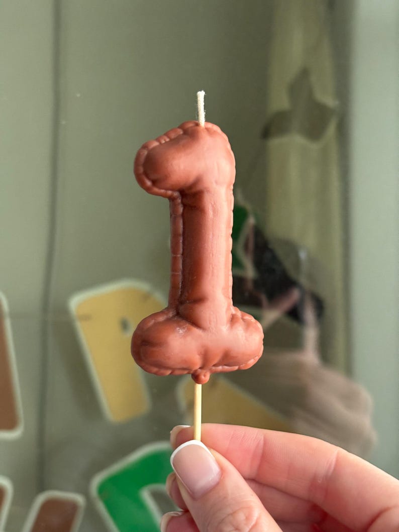 May include: A brown, number one shaped candle with a smooth, matte finish and stitched-like detailing along the edges. The candle is mounted on a thin, light-colored wooden stick and features a single white wick protruding from the top center.