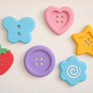 May include: A collection of six colorful buttons in various shapes including a light blue butterfly, a pink heart, a yellow star, a red strawberry with a green top, a purple circle, and a light blue flower with a white spiral center design.