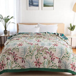 Puede incluir: Un edredón estampado con flores blancas y verdes con un borde verde oscuro. El edredón está doblado en una cama con almohadas blancas.