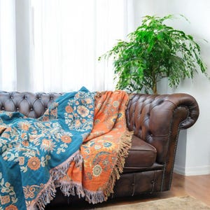 May include: A brown leather sofa with a teal and orange patterned throw blanket. The sofa is in a living room with a large green plant in the background.