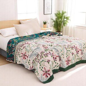 May include: A floral patterned throw blanket with a green border. The blanket is folded on a bed with white bedding and pillows.