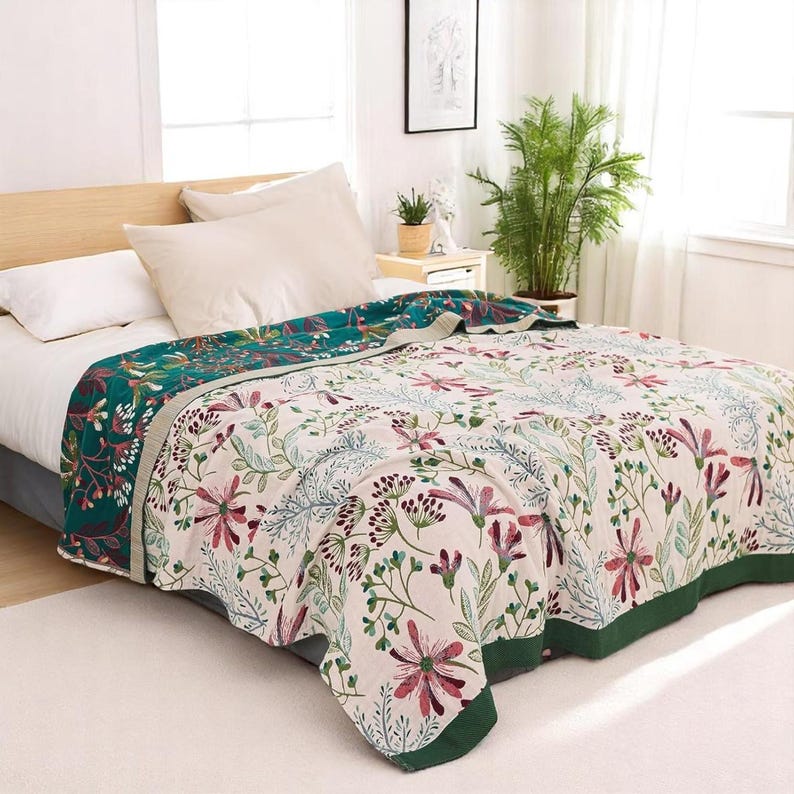 May include: A floral patterned throw blanket with a green border. The blanket is folded on a bed with white bedding and pillows.