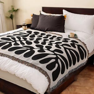 Puede incluir: Una manta tejida en blanco y negro con un patrón de hojas. La manta está colocada sobre una cama blanca con almohadas blancas y negras.