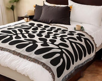 May include: A black and white woven throw blanket with a leaf pattern. The blanket is draped over a white bed with black and white pillows.