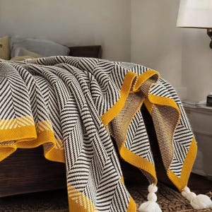 Könnte beinhalten: Eine schwarze und weiße Chevron-Muster-Wurfdecke mit gelbem Rand und weißen Quasten. Die Decke ist gefaltet und über ein Bett gelegt.