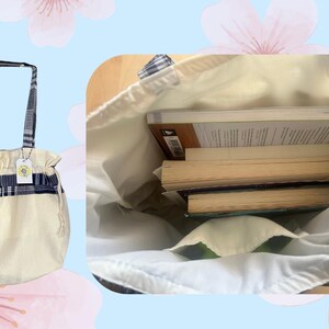 May include: A white canvas tote bag with blue and white plaid trim. The bag has two straps and a drawstring closure. The bag is filled with three books.