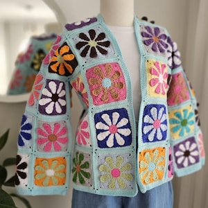 Könnte beinhalten: Ein offener Häkelcardigan mit quadratischen Blumenmustern. Die Blumen sind in verschiedenen Farben wie Rosa, Gelb, Orange und Weiß auf hellblauem Hintergrund gehalten. Der Cardigan wird auf einer Schaufensterpuppe präsentiert, was seine Handarbeit hervorhebt.