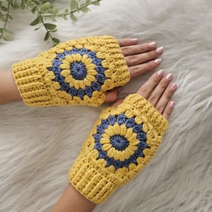 May include: A pair of yellow crochet fingerless gloves with a blue and yellow floral design. The gloves are handmade and feature a ribbed cuff. The gloves are displayed on a white, fluffy surface.