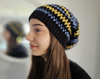 May include: A black knit beanie with a zig-zag pattern in yellow and light blue. The hat is worn by a person with long brown hair. The beanie is a stylish accessory for cold weather.