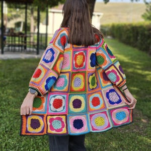 May include: A colorful, handmade crochet cardigan. The long-sleeved garment features a patchwork design of squares in various bright colors, including red, blue, yellow, and green. The cardigan is knee-length and has a black border.