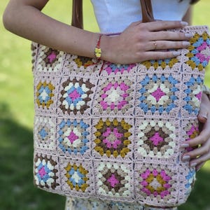 Könnte beinhalten: Handgefertigte Häkeltasche mit Patchwork-Design. Die Tasche ist hauptsächlich hellrosa mit farbigen Quadraten in Blau, Rosa, Braun und Gelb. Sie hat braune Ledergriffe und eine strukturierte, offene Webart.