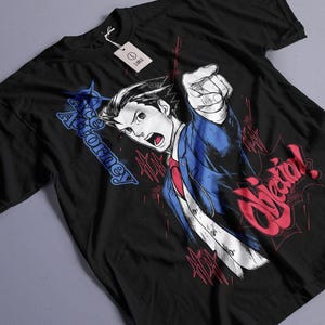 May include: A black t-shirt featuring a graphic of a character pointing forward, wearing a blue suit and red tie. The text "Ace Attorney" appears in blue, and the word "Objection!" is printed in large red letters. A size "L" tag is attached.