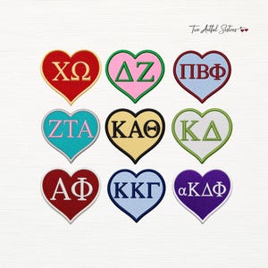 May include: Nine heart-shaped embroidered patches arranged in a grid. Each patch features Greek letters in various colors including red, pink, light blue, turquoise, yellow, white, and purple, with contrasting borders and the text Two Artful Sisters.