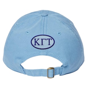 May include: Pale blue baseball cap with an embroidered navy blue oval patch displaying the letters "K&Gamma;T". The cap features a curved peak, adjustable strap, and a metal buckle.