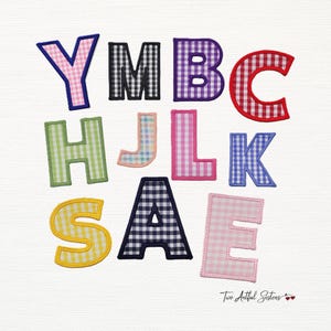 May include: Assorted fabric letters in various colors and gingham patterns, including pink, black, purple, red, green, blue, and yellow. The letters are outlined with contrasting stitching. The image includes the text "Two Artful Sisters".