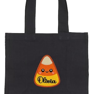 Peut inclure: Sac fourre-tout noir avec un motif de bonbon en forme de ma&iuml;s. Le bonbon est orange, jaune et blanc avec un visage souriant et le nom "Olivia" brod&eacute; dessus. Le sac a deux poign&eacute;es noires.