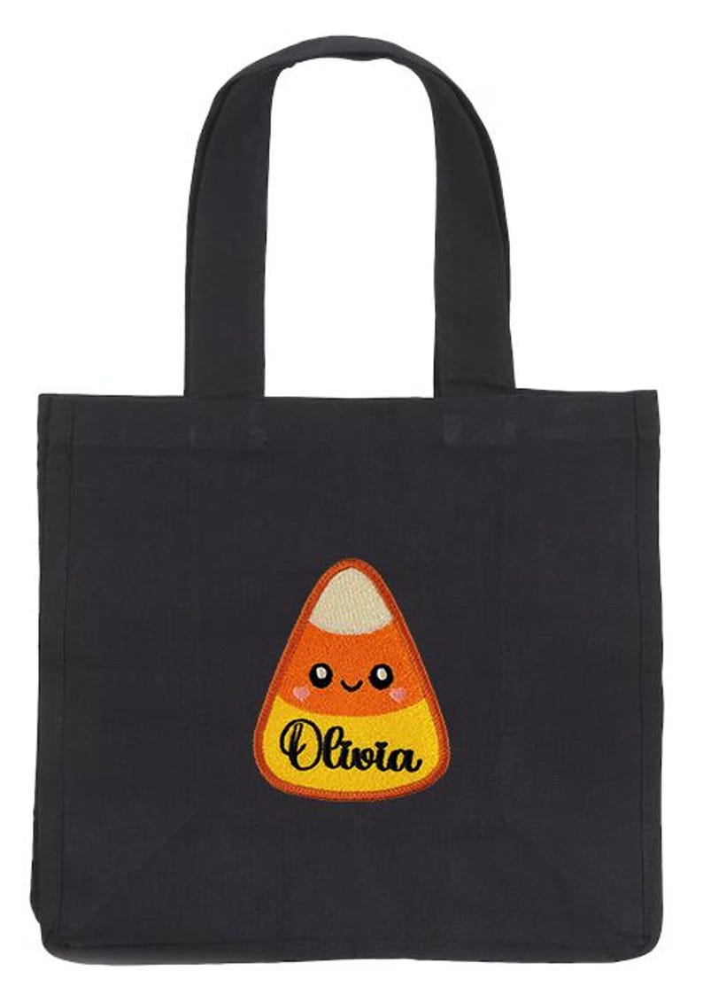 Peut inclure: Sac fourre-tout noir avec un motif de bonbon en forme de ma&iuml;s. Le bonbon est orange, jaune et blanc avec un visage souriant et le nom "Olivia" brod&eacute; dessus. Le sac a deux poign&eacute;es noires.