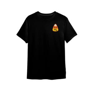 Peut inclure: T-shirt noir avec un motif de bonbon en forme de ma&iuml;s et le nom "Betty". Le motif de bonbon comprend des couleurs orange, blanc et jaune. Le t-shirt est &agrave; manches courtes et &agrave; col rond.