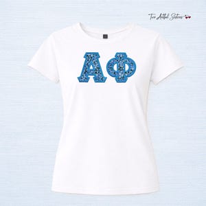 May include: White t-shirt featuring the Greek letters ΑΦ in a blue floral pattern. The letters are outlined in a darker blue. The shirt has short sleeves and a crew neck. The text "Two Artful Sisters" is in the upper right corner.