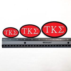 May include: Three red oval patches with the Greek letters TKS in white, each with a black border. A ruler with inch measurements is below the patches, showing their varying sizes.