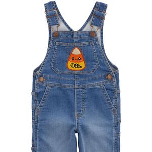 Peut inclure: Salopette en jean bleu avec bretelles r&eacute;glables et boutons m&eacute;talliques. Un &eacute;cusson de bonbon en forme de ma&iuml;s avec le nom "Ollie" est cousu sur la bavette. La salopette a des poches et est con&ccedil;ue pour les enfants.