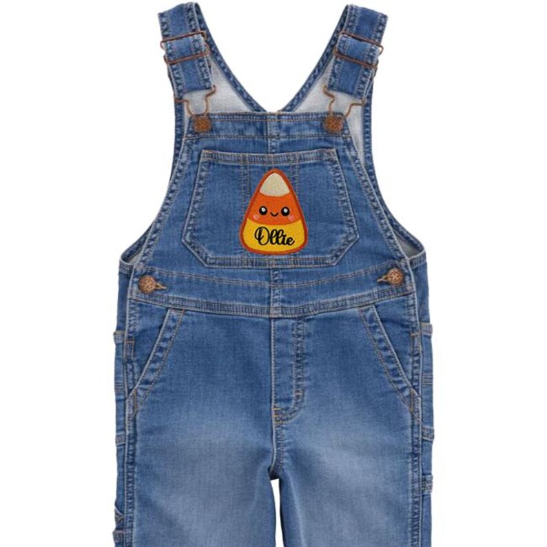 Peut inclure: Salopette en jean bleu avec bretelles r&eacute;glables et boutons m&eacute;talliques. Un &eacute;cusson de bonbon en forme de ma&iuml;s avec le nom "Ollie" est cousu sur la bavette. La salopette a des poches et est con&ccedil;ue pour les enfants.