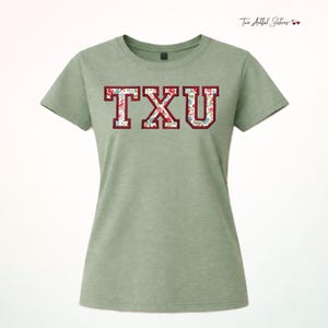 May include: A sage green short-sleeved t-shirt featuring the letters TXU in a collegiate block font. The letters have a dark red outline and are filled with a floral pattern consisting of small red, blue, and white flowers on a white background.