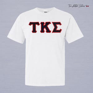May include: A white short-sleeve crew neck t-shirt featuring the Greek letters TKE in black with a red outline centered on the chest. The shirt has a standard fit with a ribbed collar and a small brand tag visible at the inner neckline.