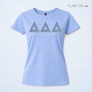 May include: A light blue short-sleeve t-shirt featuring three embroidered Greek delta symbols across the chest. The triangles are outlined in a metallic gold thread with a light blue fill. The shirt has a crew neckline and a heathered fabric texture.