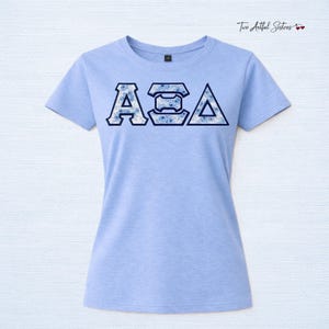 May include: A light blue short-sleeved t-shirt featuring the Greek letters Alpha Sigma Alpha in a dark blue outline. The letters are filled with a white and blue floral pattern. The text Two Artful Sisters is visible in the top right corner.