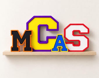 Könnte beinhalten: Ein Set aus vier freistehenden Blockbuchstaben, die das Wort MCAS bilden. Das M ist schwarz mit orangefarbenem Rand, das C ist gelb mit violettem Rand, das A ist blau mit gelbem Rand und das S ist weiß mit rotem Rand, auf einem Regal.