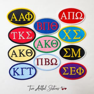 May include: A collection of embroidered oval patches in various colors, each featuring Greek letters. The patches have a stitched border and are in colors like black, red, blue, yellow, and pink. The text "Two Artful Sisters" is at the bottom.