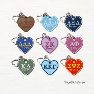 May include: Heart-shaped keychains in various colors, each featuring Greek letters and phrases like "Alumna," "Little Sis," and "Forever." The keychains have silver-tone clasps and are from "Two Artful Sisters."