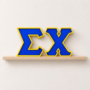 May include: A three-dimensional Greek letter Sigma and Chi symbol set. Each letter features a vibrant blue center face with a thick, raised yellow border, creating a bold contrast. The letters are positioned side-by-side on a flat surface.