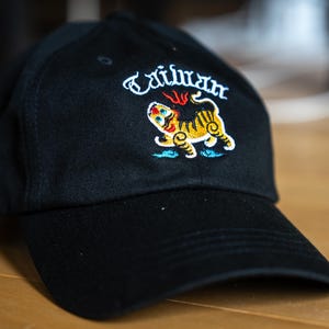 May include: Black baseball cap with embroidered design. The design features the word "Taiwan" in white script above a colorful tiger illustration in yellow, orange, and blue. The cap has a curved brim.