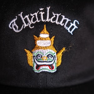 Può includere: Cappellino da baseball nero con la scritta "Thailand" ricamata sopra un'immagine colorata e dettagliata di una figura tradizionale thailandese. La figura ha un copricapo dorato, un viso verde e una bocca rossa.
