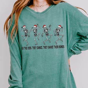 May include: Light green long-sleeve shirt with a crew neck. The shirt features a graphic of four dancing skeletons wearing Santa hats, with the text "SO THE KIDS, THEY DANCE, THEY SHAKE THEIR BONES".