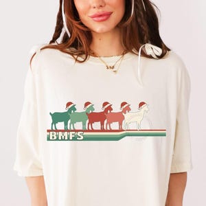 Può includere: T-shirt color crema con una grafica di sei capre in fila, ognuna con un cappello da Babbo Natale. Le capre sono in tonalità di verde, rosso e bianco, con il testo "BMFS" sotto. Un design a strisce rosse e verdi è sotto le capre.