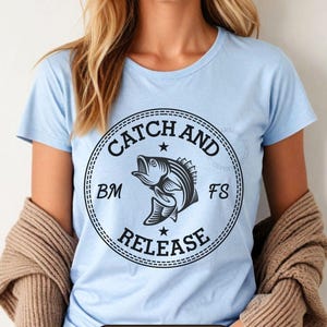 Op de afbeelding: Lichtblauw T-shirt met een cirkelvormige afbeelding met de tekst "Catch and Release" en een illustratie van een vis. Het shirt heeft ook de tekst "Light Blue" aan de onderkant. Een vrouw draagt het shirt.