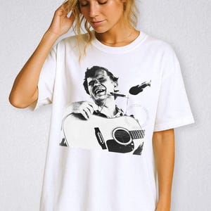 Può includere: T-shirt bianca con un'immagine in bianco e nero di un musicista che suona la chitarra e l'armonica. La grafica è un ritratto di un uomo con una chitarra. La maglietta ha maniche corte e girocollo.