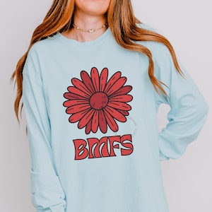 May include: Pale blue long-sleeved shirt featuring a large red daisy design and the letters "BMFS" in red. The shirt also has the word "CHAMBRAY" in a brown rectangle at the bottom.